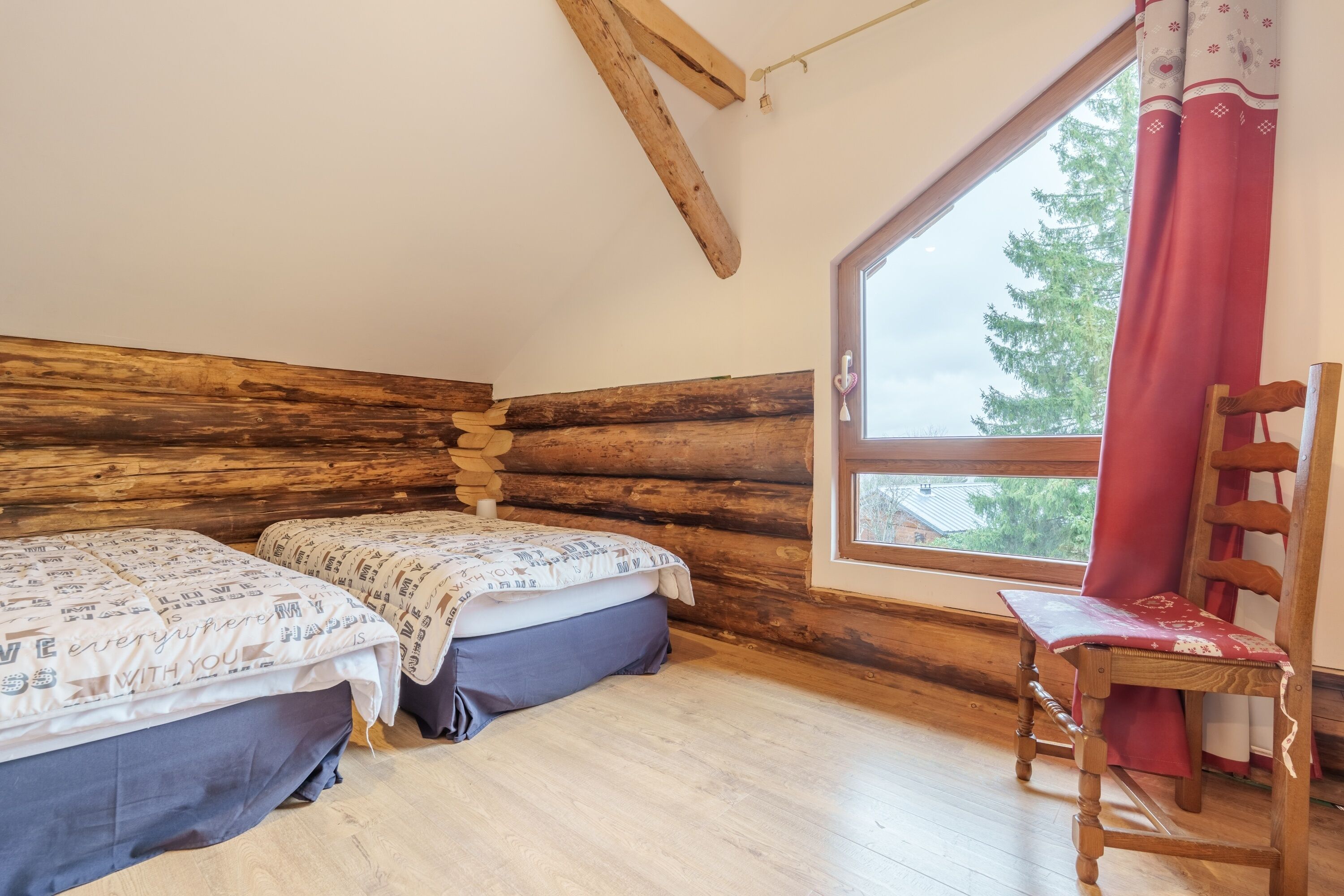 Habitación tipo cabaña de madera con dos camas, una silla de madera y un ventanal con vistas a árboles.