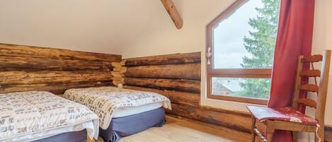 Habitación tipo cabaña de madera con dos camas, una silla de madera y un ventanal con vistas a árboles.