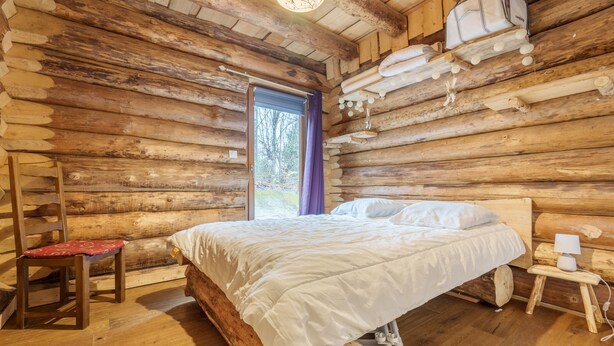 Quarto de cabana rústica com uma cama, uma cadeira, uma mesinha com abajur e uma janela com cortina.