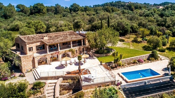 Una casa de piedra de dos pisos con piscina, rodeada de exuberante vegetación y colinas.