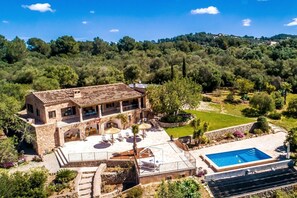 Uma casa de pedra de dois andares com piscina, rodeada por vegetação exuberante e colinas.