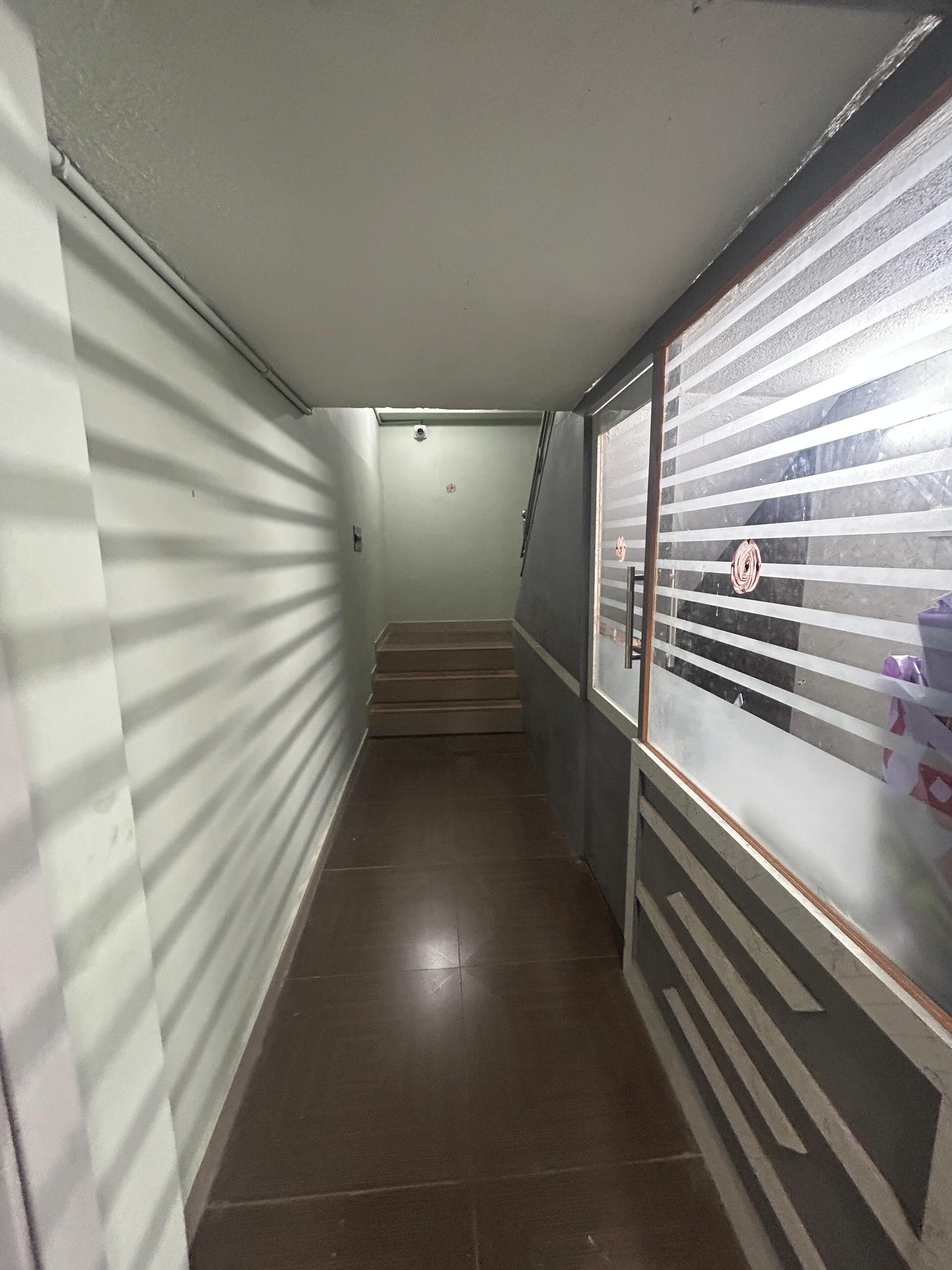 A narrow hallway with a staircase leading to an upper floor. The walls are plain with a light color, and there is a window with horizontal blinds on the right.