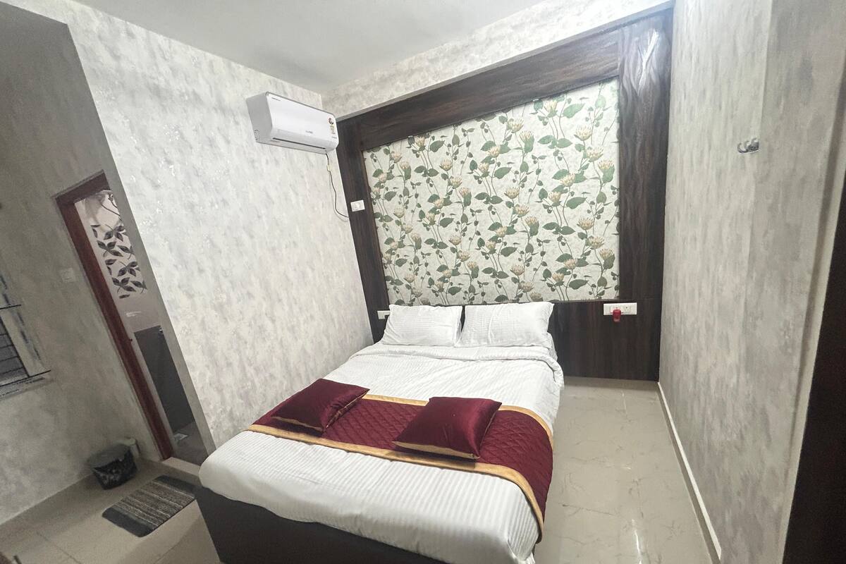 Une chambre à coucher avec un lit, une fenêtre ornée de rideaux à motifs floraux, un climatiseur fixé au mur et une porte décorée d’un motif ornemental.