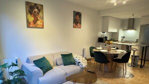 En moderne stue med en sofa, et sofabord i glas og et spiseområde med stole og bord. I baggrunden er der et køkken med en mikrobølgeovn og et køleskab.