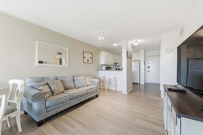 A modern living room with a grey sofa, white chairs, a kitchen area with white cabinets, and a flat-screen TV. - Coastal 2BR Condo w Ocean Views & Pool (Hilton Head Island)