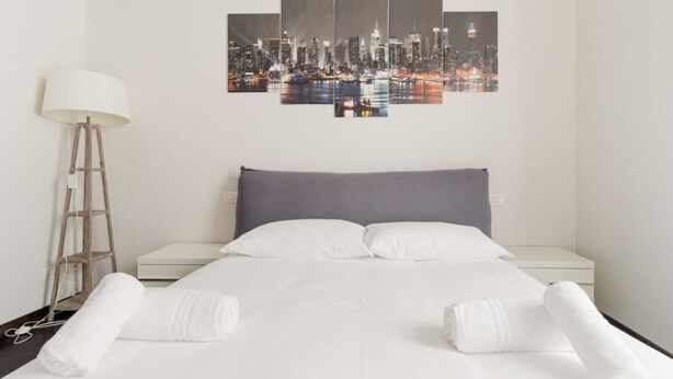 A hotel room with a bed, white linens, a cityscape artwork, a lamp, and a nightstand. - GuestHost - Cozy 50m2 apartment located on the second floor of a building with lift, ideal for 4 people.The area is well equipped with the main services and allows you to quickly reach the most important attractions thanks to the presence of various bus, (Milan)