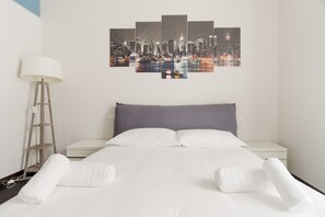 Une chambre d’hôtel avec un lit, du linge de lit blanc, une œuvre d’art représentant un paysage urbain, une lampe et une table de chevet.