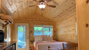 Een knus vakantiehuisje met houten inrichting, een bed met witte lakens, een eettafel en een plafondventilator.
