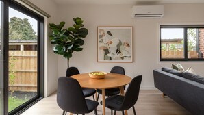 Ett modernt matområde med ett runt bord, svarta stolar och en skål med frukt. Här finns också en stor växt, en inramad abstrakt målning och en grå soffa.