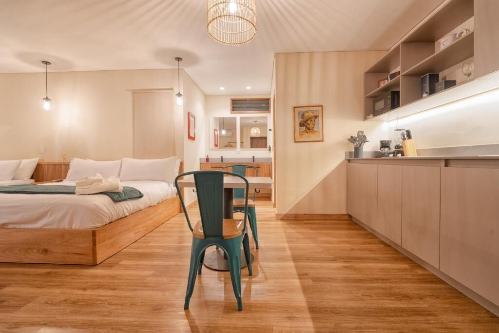 A modern hotel room with a wooden bed, a dining table with chairs, a kitchen area, and a bathroom visible through an open door.