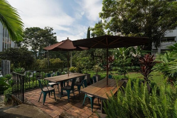 An outdoor patio with a wooden table and chairs, surrounded by lush greenery and a large umbrella. - Candelaria Aparta Hotel Boutique  (Medellín)