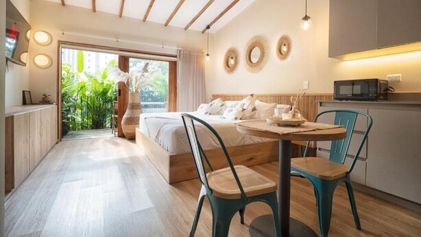 Een moderne slaapkamer met een bed, een eettafel en stoelen. Er is een keukenhoek met een magnetron en kastjes. De kamer heeft een houten vloer en uitzicht op groen.