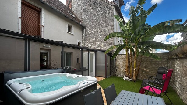 A hot tub in a backyard with a table and chairs, surrounded by a stone wall and a banana tree. - Charming 6-person home with garden and jacuzzi in La Roche-Posay (La Roche-Posay)