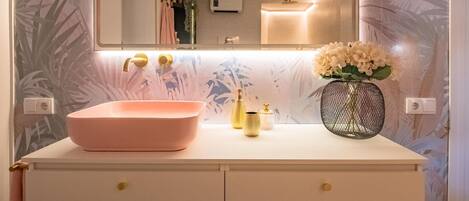 Un baño con un lavamanos rosa, detalles dorados y un florero con flores.