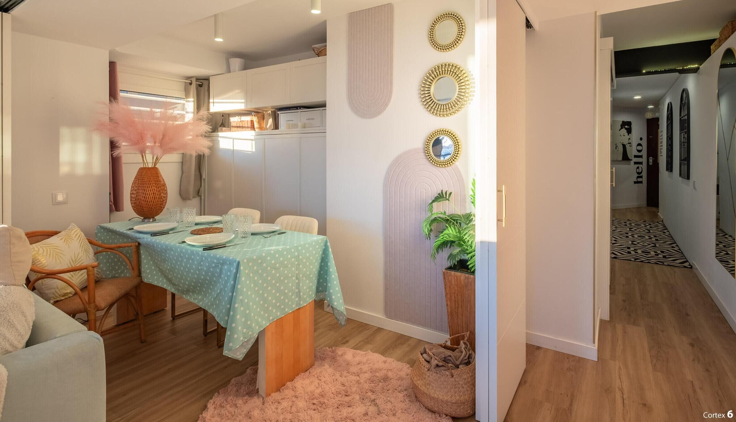 Un coin repas avec une table dressée pour quatre personnes, une cuisine en arrière-plan et un couloir agrémenté d’une pancarte indiquant « Bonjour ».