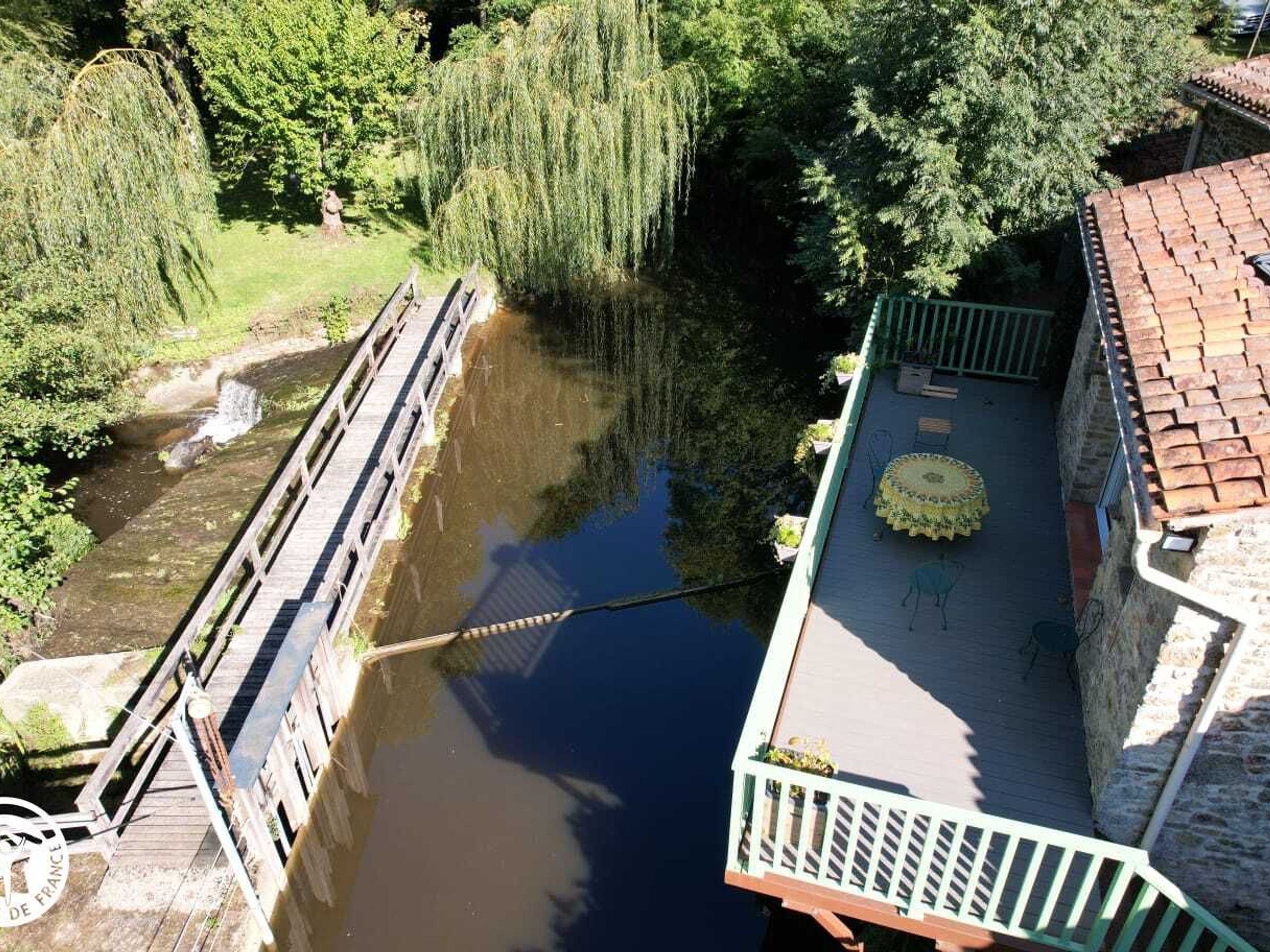 Une promenade au bord de la rivière, avec un pont en bois, un bâtiment en pierre doté d’un balcon et une végétation luxuriante.