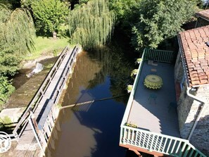 A riverside walkway with a wooden bridge, a stone building with a balcony, and lush greenery.