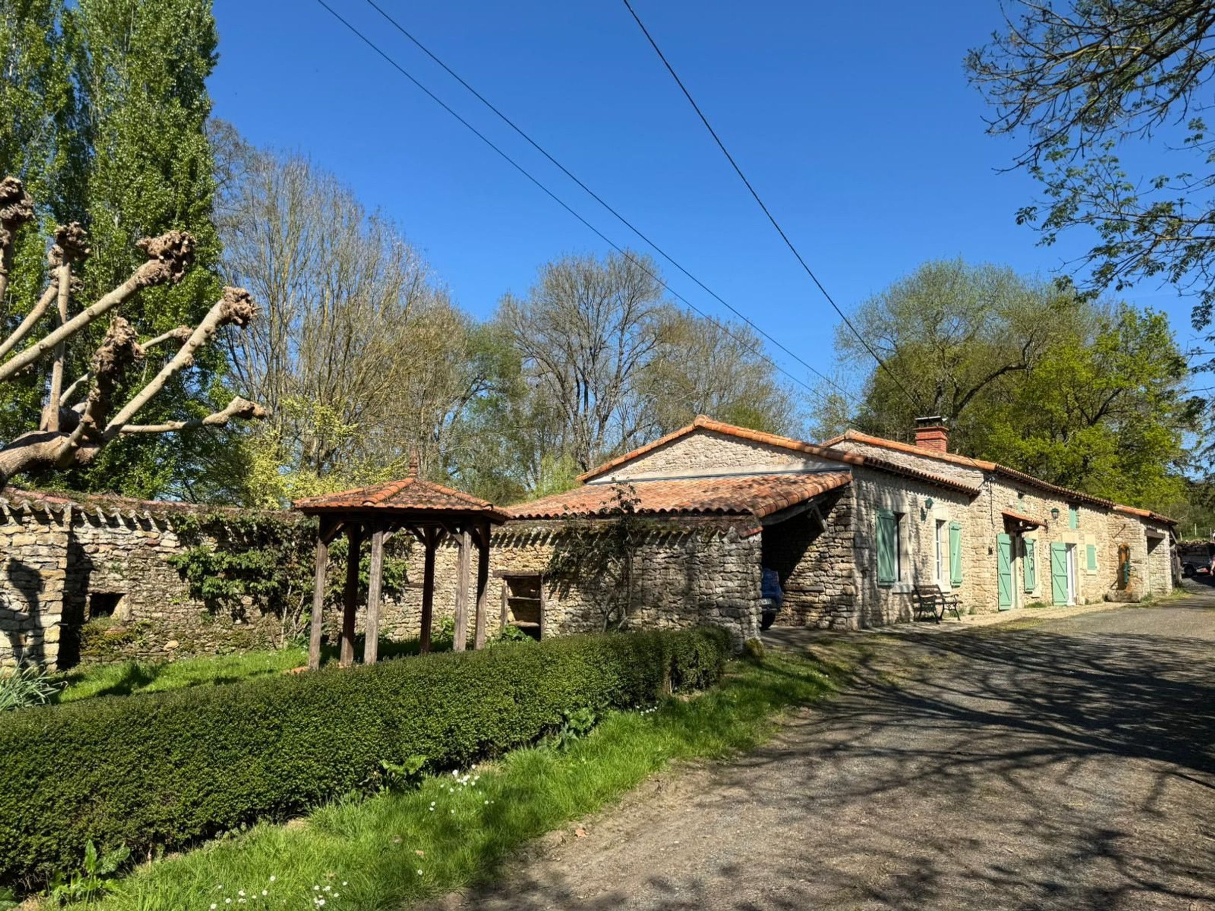 A stone building with a tiled roof, a wooden structure with a roof, and a well-maintained hedge.