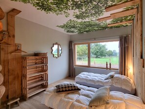 A cozy bedroom with a large window, wooden furniture, and a view of a green landscape. - Cozy Lodge in Vivans with Pool and Spa, Pets Allowed (Vivans)