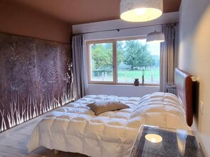 Une chambre avec un grand lit, une fenêtre donnant sur un paysage verdoyant et un luminaire suspendu moderne.