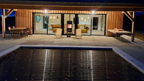 A modern wooden cabin with a large sliding glass door entrance, a fireplace, and a patio area with outdoor seating. - Cozy Lodge in Vivans with Pool and Spa, Pets Allowed (Vivans)