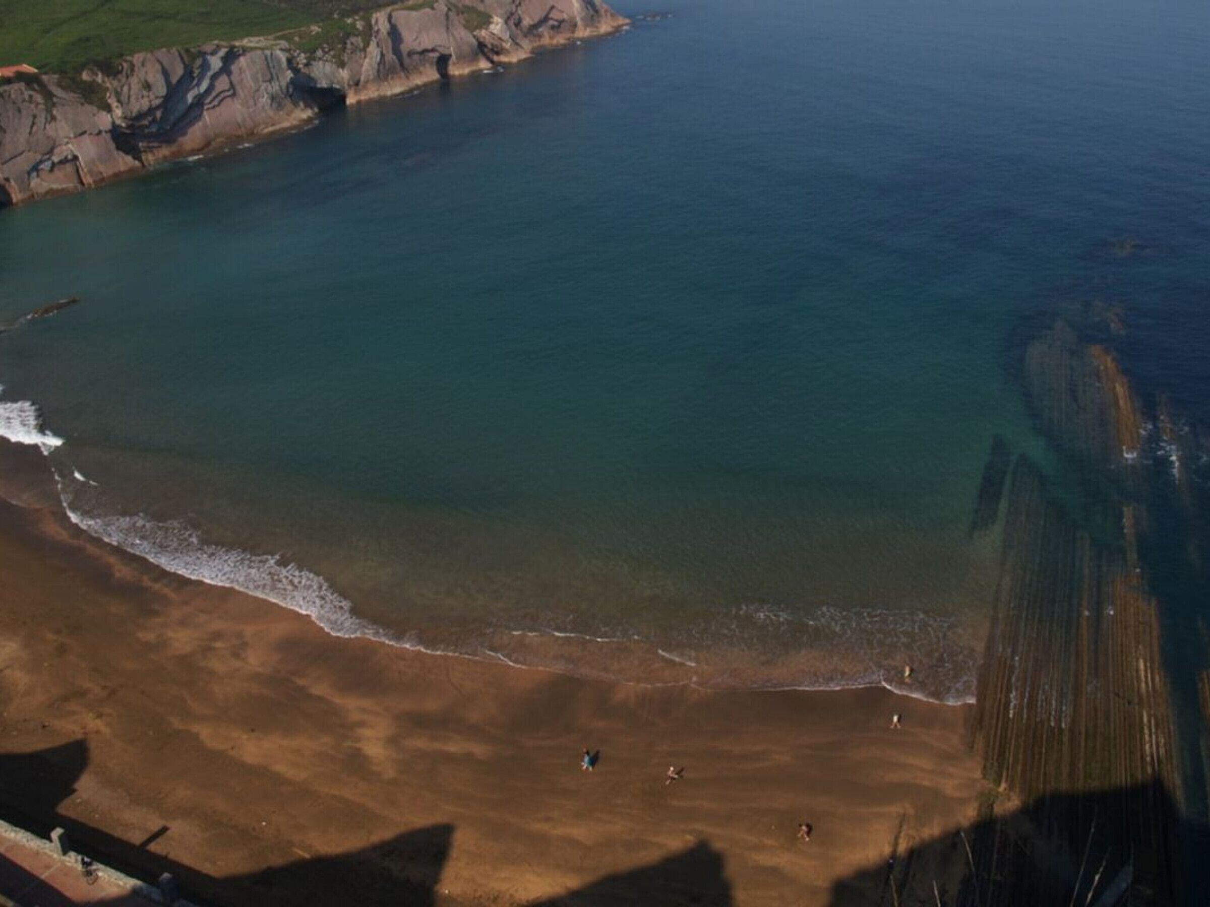 Vista aérea de una playa con aguas cristalinas, acantilados rocosos y algunas personas caminando por la orilla.