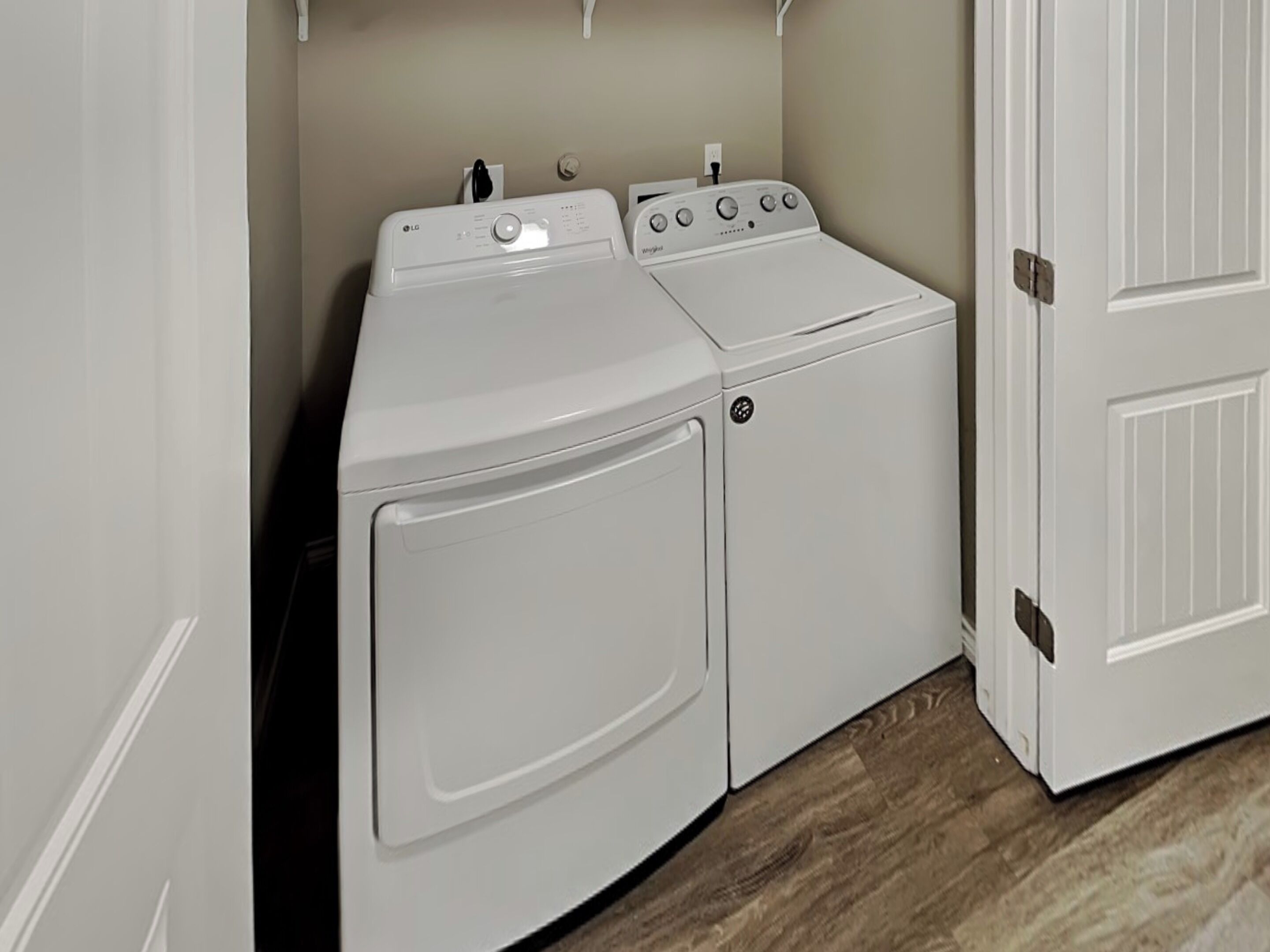 A laundry room with a front-loading washer and dryer, a white door, and a wooden floor.