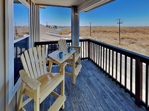 Eine Holzterrasse mit zwei Adirondack-Stühlen und einem Tisch, mit Blick auf eine flache Landschaft, ein Gewässer und Stromleitungen in der Ferne.
