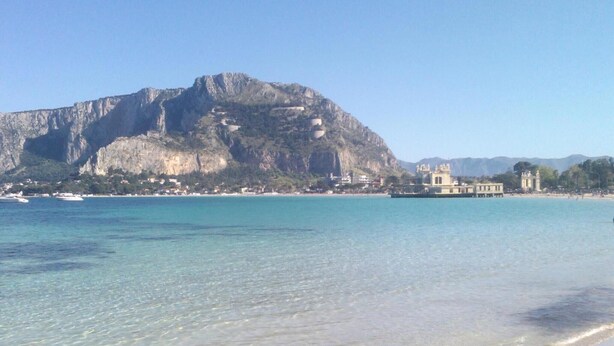 Ein Küstenstrand mit klarem Wasser, ein markanter Berg im Hintergrund und ein Gebäude im klassizistischen Stil.
