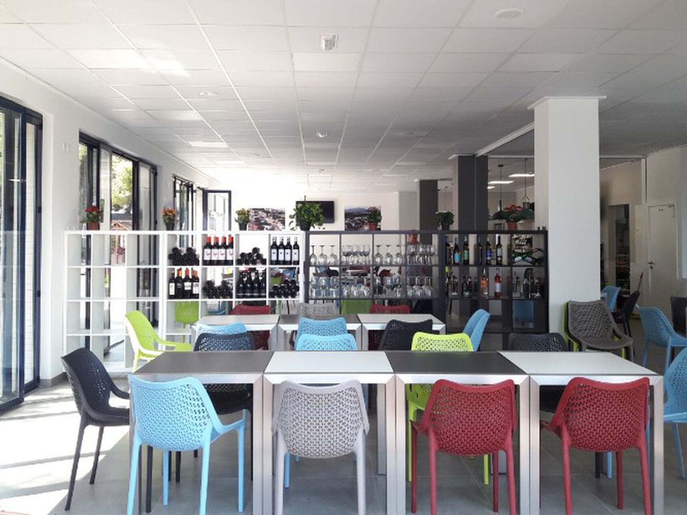 A modern indoor space with tables and chairs, shelves with bottles and glasses, and large windows allowing natural light.
