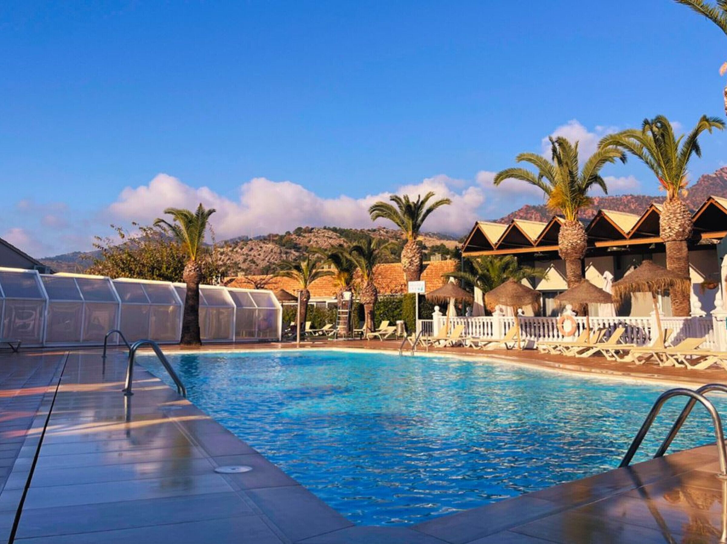 Un espace piscine avec une eau d’un bleu limpide, des palmiers et des bungalows sur pilotis.