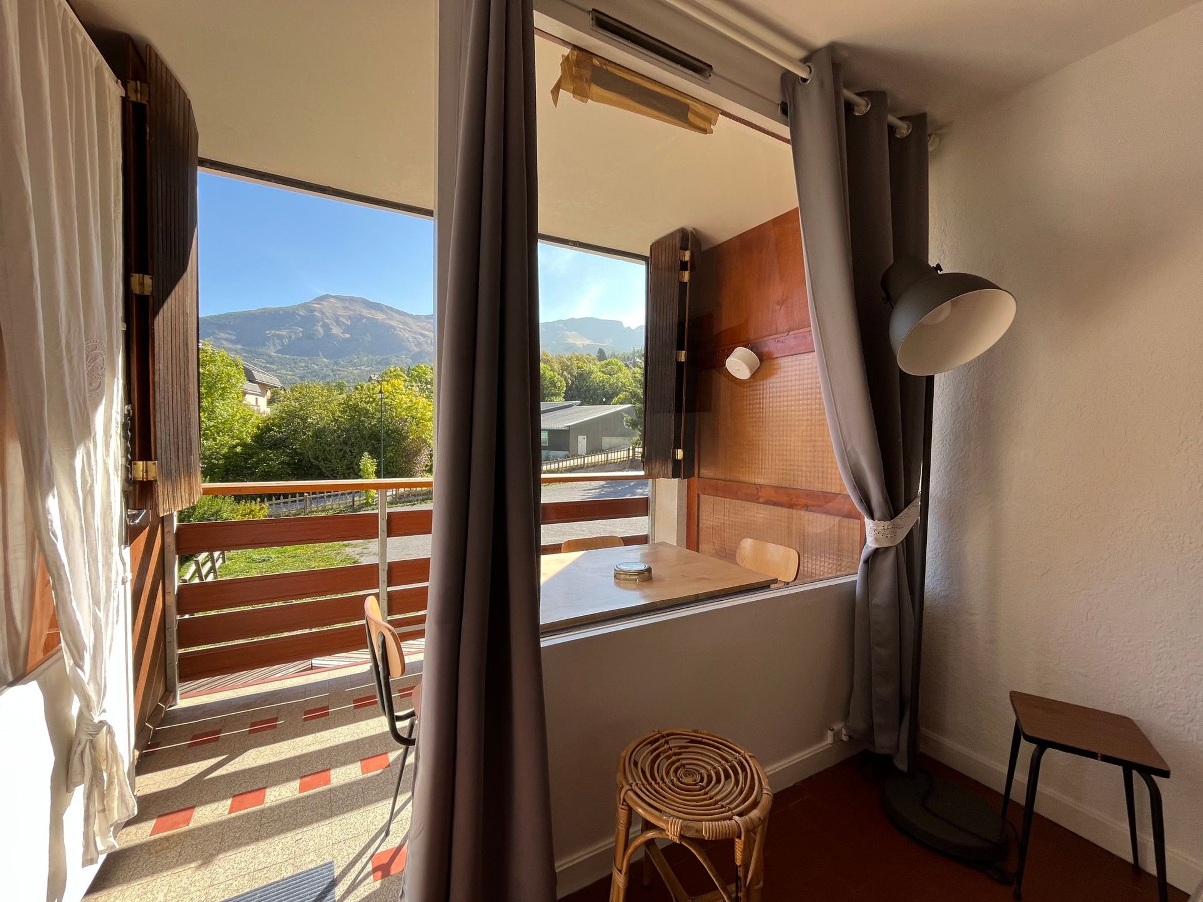 A room with a view of mountains, a wooden chair, a small table, and a window with curtains.