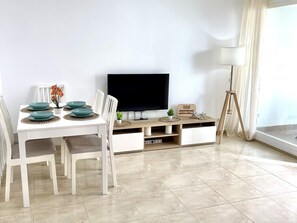 Un saló modern amb taula de dinar blanca per a quatre, televisió plana penjada a la paret, un moble d'aixetes de fusta amb espais d'emmagatzematge i una finestra gran amb cortines.