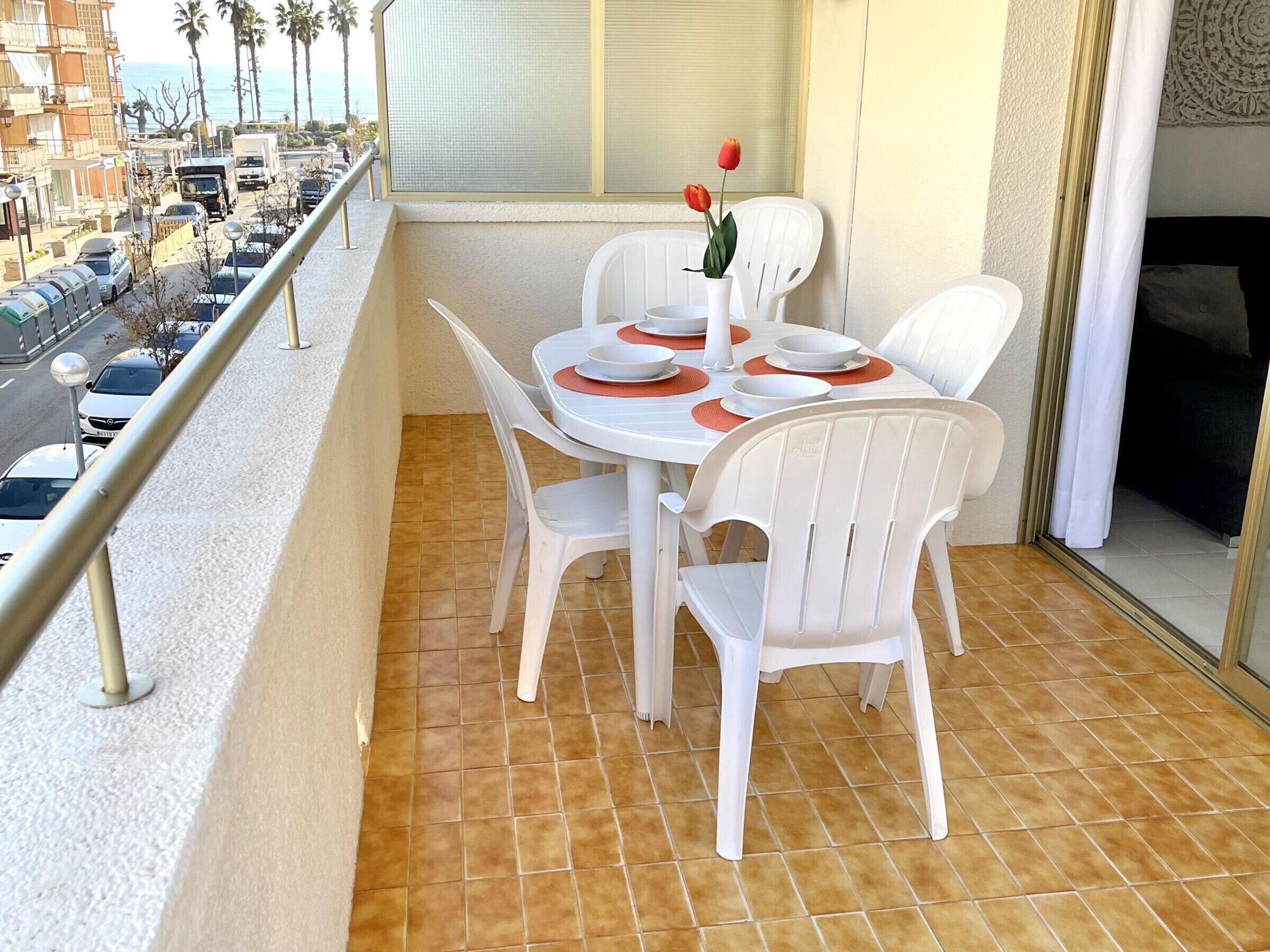 Een balkon met een tafel gedekt voor twee personen, witte plastic stoelen, een vaas met een enkele rode bloem, en uitzicht op een straat met geparkeerde auto's en palmbomen.