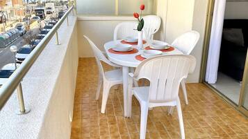 Een balkon met een tafel gedekt voor twee personen, witte plastic stoelen, een vaas met een enkele rode bloem, en uitzicht op een straat met geparkeerde auto's en palmbomen.