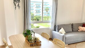 Un salon moderne avec une table à manger en bois, des chaises en osier, un canapé gris et une vue sur un immeuble d’habitation à travers de grandes fenêtres.