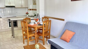 A modern kitchen with white cabinets, a dining table with chairs, a grey sofa with an orange pillow, and a wooden coffee table with a plant. - Vancouver Relax: Air-Conditioned Apartment with Pool, Parking, and 7-Min Walk to Cala Fon Beach (Salou)