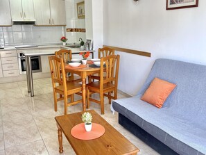 A modern kitchen with white cabinets, a dining table with chairs, a grey sofa with an orange pillow, and a wooden coffee table with a plant. - Vancouver Relax: Air-Conditioned Apartment with Pool, Parking, and 7-Min Walk to Cala Fon Beach (Salou)
