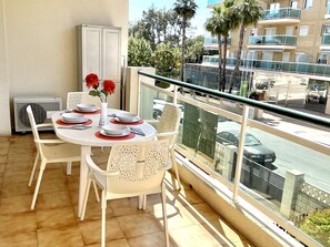 A balcony with a dining table set for four, white chairs, a white cabinet, and a view of a street with palm trees and buildings. - Vancouver Relax: Air-Conditioned Apartment with Pool, Parking, and 7-Min Walk to Cala Fon Beach (Salou)