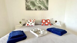 Una cama bien hecha con cubrecolcha gris texturizado, dos toallas azules, una bandeja con taza y platillo, y dos almohadas con diseños geométricos.