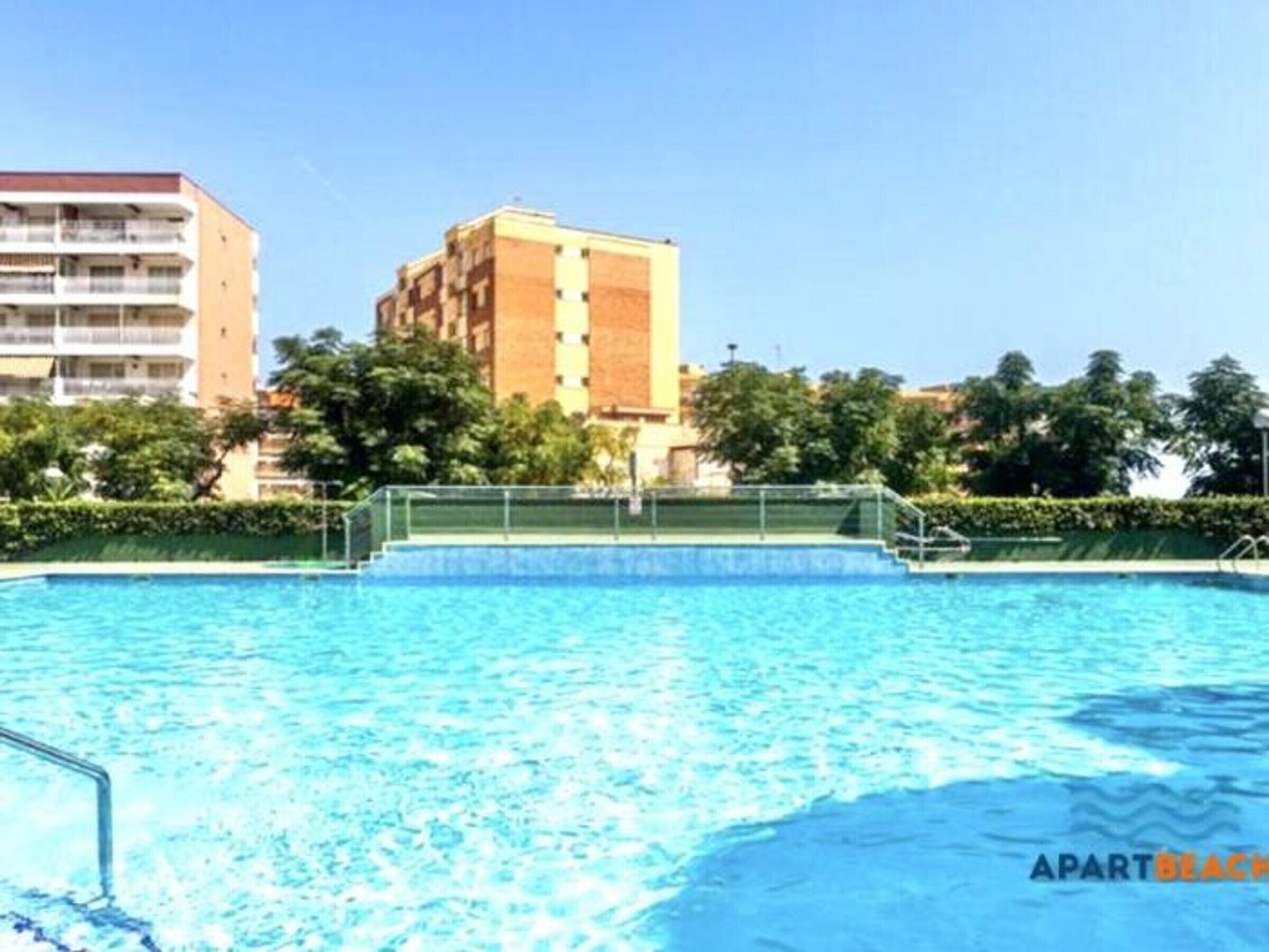 A swimming pool with a clear blue water surface, surrounded by a metal fence and a staircase leading down to the pool. In the background, there are multi-story buildings and lush green trees.