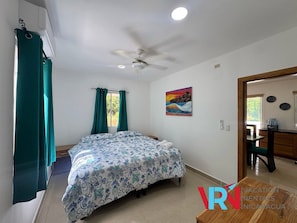 A bedroom with a bed, a ceiling fan, a window with curtains, a painting, and a view of the outside.