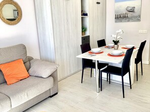 Un saló modern amb un sofà gris, coixins taronja, una taula de dinar posta amb plats i coberts, i un quadre d'una barca a la paret.