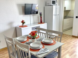 Ein Essbereich mit einem weißen Tisch, gedeckt für vier Personen, orangefarbenen Platzsets, weißen Tellern und einer roten Vase mit Blumen. Im Hintergrund ist ein weißes Schränkchen mit einer Mikrowelle darauf zu sehen.