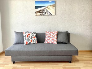 Ein graues Sofa mit drei Kissen vor einer strukturierten Wand, darüber ein gerahmtes Bild und ein Holzboden.