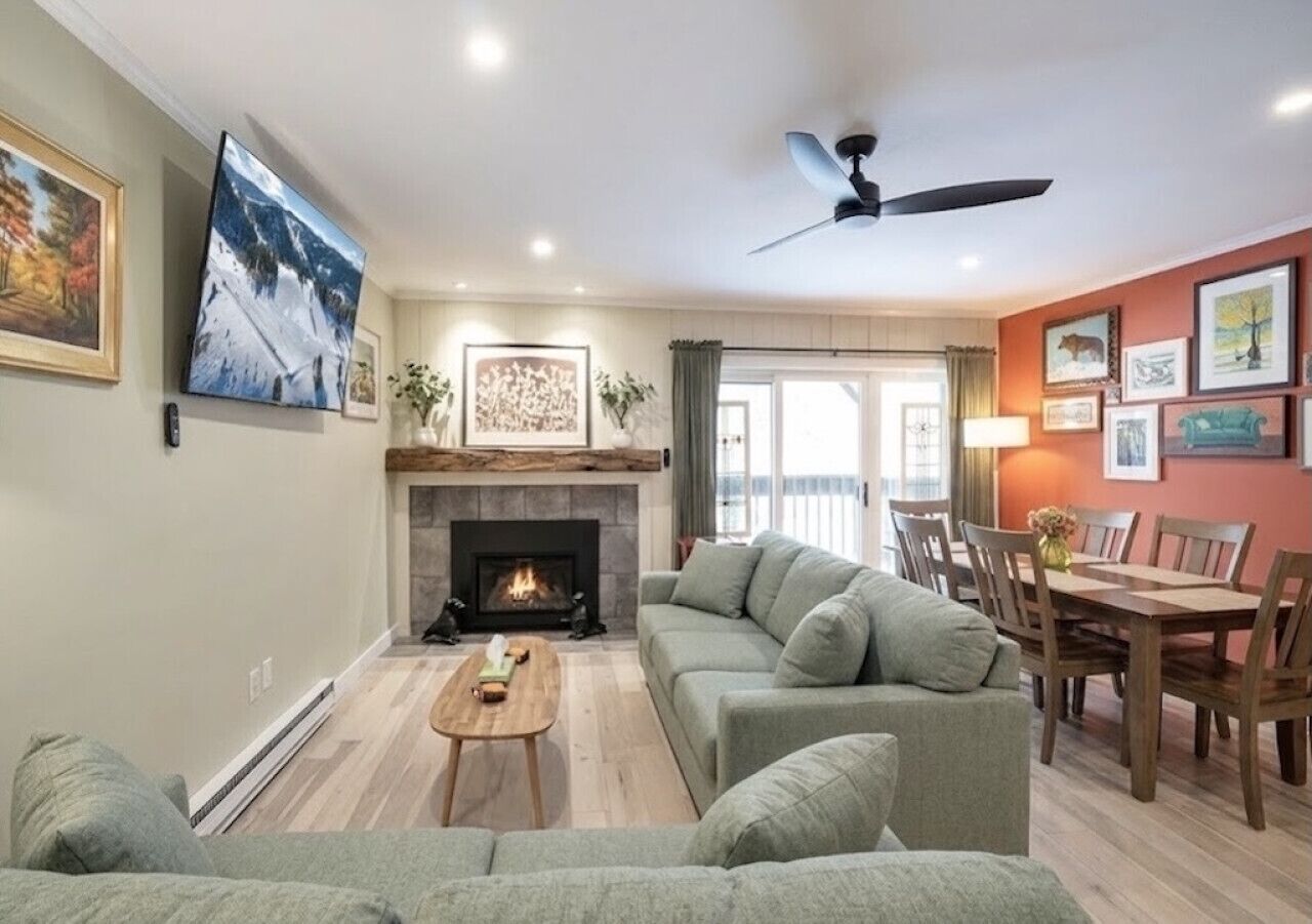 A living room with a sofa, a coffee table, a fireplace, a dining area with chairs, and a ceiling fan.