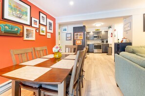 A dining area with a wooden table, chairs, and a vase of flowers. There is a kitchen in the background with modern appliances.