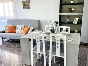 Ein modernes Wohnzimmer mit grauem Sofa, weißen Sesseln, einem Esstisch für vier Personen und einem Bücherregal mit Dekoration.
