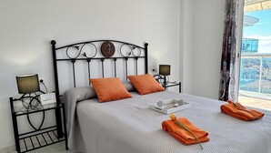 Una cama bien hecha con una colcha gris texturizada, dos almohadas naranjas y una bandeja con tazas y una tetera a un lado.