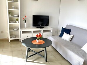 Una sala de estar moderna con una mesa negra redonda, un sofá gris con cojines azules, un mueble blanco para televisor con una TV de pantalla plana y un estante blanco con objetos decorativos.